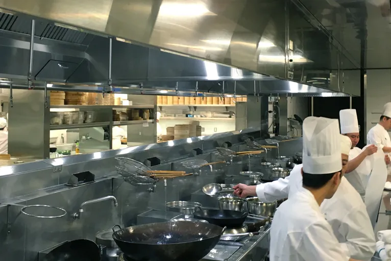 Professional chefs cooking in a commercial kitchen with large overhead ventilation systems, showing integration of AC unit in a commercial kitchen for heat management.